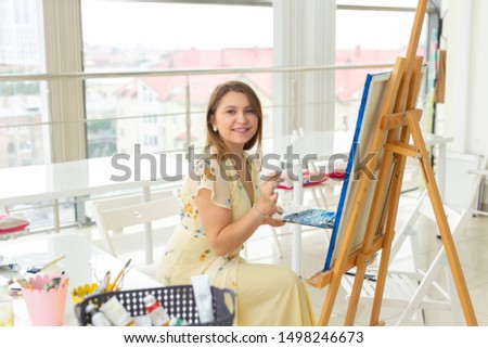 Art school, creativity and leisure concept - student girl or young woman artist with easel, palette and paint brush painting picture at studio