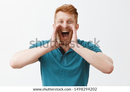 Carefree friendly redhead caucasian man in green polo shirt holding palms near opened mouth while yelling on distance, trying call friend who walking far away, shouting out loud over gray background