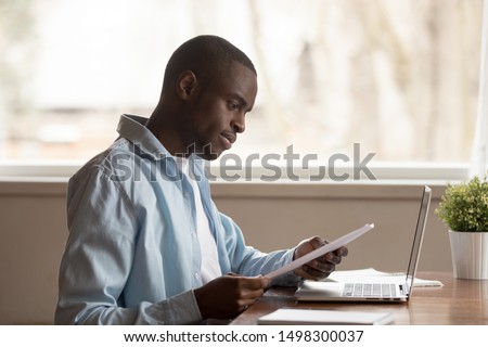 Focused african American millennial man sit at table work study using laptop reading paper document, concentrated biracial male look through consider paperwork letter, good news notice at workplace Royalty-Free Stock Photo #1498300037