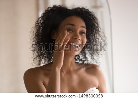 Smiling african American young woman wrapped in towel after shower look in mirror apply under eye anti-aging cream, happy biracial female perform morning skincare beauty procedures at home Royalty-Free Stock Photo #1498300058