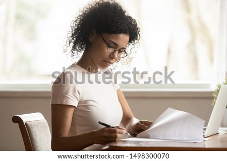 Focused mixed race ethnicity millennial businesswoman in glasses work at laptop read sign paper documents, concentrated african American woman student consider paperwork do corrections Royalty-Free Stock Photo #1498300070