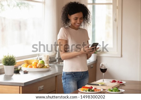 Happy african American millennial woman chop slice vegetables drink wine cooking in kitchen using cellphone, smiling biracial female hold smartphone texting messaging while preparing lunch