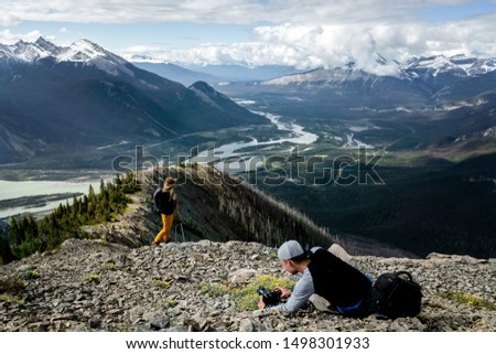Young adventure photographer taking picture of woman model in yellow trousers and brown hat hiking Greenock trail in Jasper National park, Canada. Athabasca river. Taking photos for social media