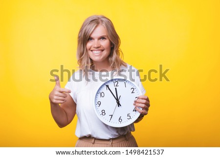 Portait of blonde woman, pointing at clock while looking at cameraover yellow wall
