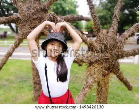 cute asian​ child girl smiling and make heart by arm and hand on top head standing on green grass in summer 