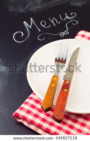 Plate, knife and fork on tablecloth over blackboard background with word menu