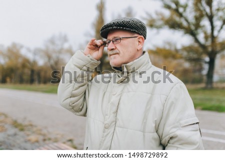 An old man in a jacket and a cap holds glasses in his hand and looks into the distance. The concept of retirement age and poor vision. Hyperopia, myopia. Royalty-Free Stock Photo #1498729892