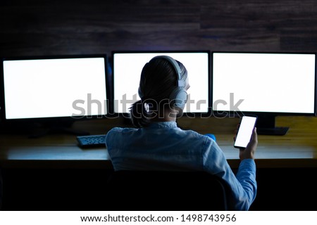 Software developer, freelancer, designer with white headphones working at workplace with wide displays and smartphone with isolated screens at night. Develops application, designs project, editing.
