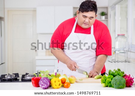Picture of Asian fat man cutting a cucumber for salad while standing in the kitchen