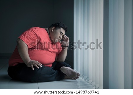 Picture of young obese man looks stressed while sitting near the window