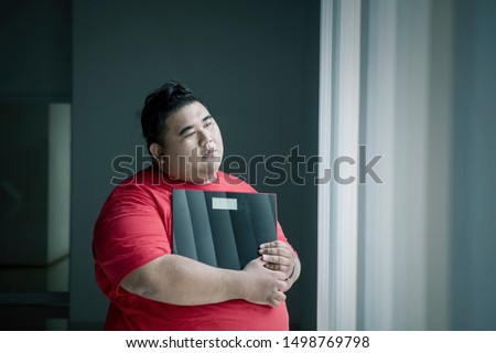 Picture of an obese man looks pensive while holding a weight scale and standing near the window