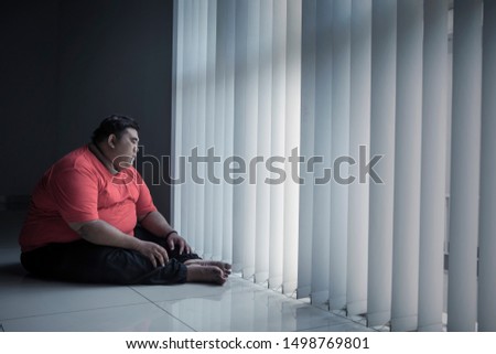 Picture of a pensive fat man looking out the window while sitting in the dark room
