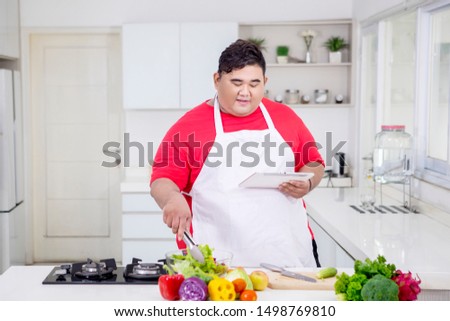 Picture of overweight man using a digital tablet while mixing salad in the kitchen