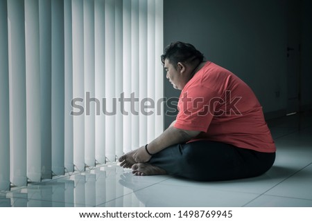 Picture of a lonely obese man looking out the window while sitting with sad expression