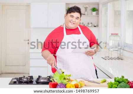 Picture of happy fat man preparing salad while using a digital tablet in the kitchen