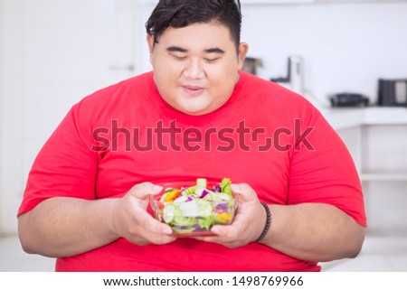Picture of young fat man looks happy while holding a bowl of tempting salad in the kitchen