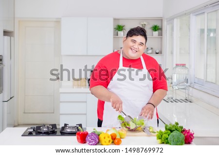 Picture of an obese man smiling at the camera while mixing fresh salad in the kitchen