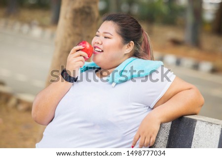Picture of happy obese woman eating fresh apple after exercise while sitting in the park