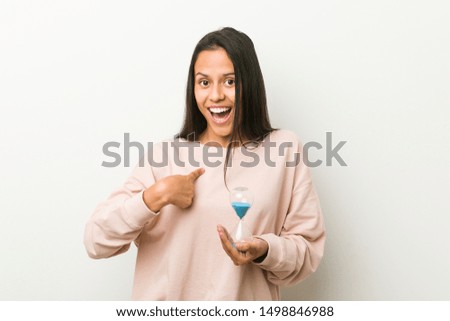 Young hispanic woman holding an hourglass surprised pointing at himself, smiling broadly.