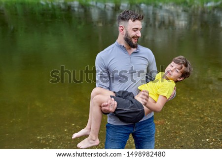 A man with a beard holds in his arms a boy of about 8 on the background of the river. Light rain on a summer day