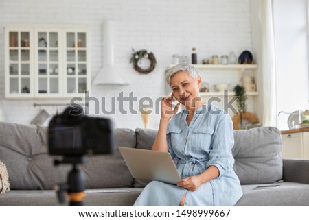 Successful senior female blogger sitting on sofa with laptop in front of camera on stand, filming broadcast, telling how to monetize video blog account. Confident mature woman is being filmed