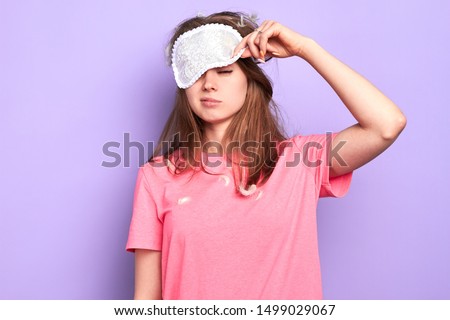 Front view of exhausted young woman raises slightly her eye mask, trying to open sleepy eyes, needs more rest after hard week, out of energy, hates morning. Sleep and wake up concept. Royalty-Free Stock Photo #1499029067