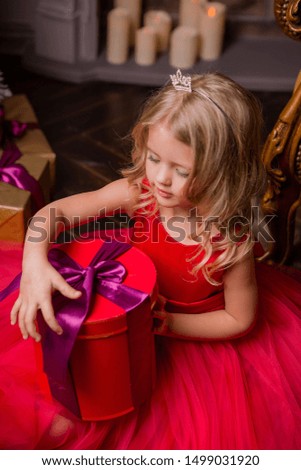 little blonde girl in evening red dress near the Christmas tree, child with Christmas gifts, the night before Christmas