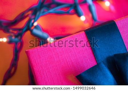 Pink yarrow box with navy blue ribbon on bright reddish background with Christmas lights. Futuristic concept.