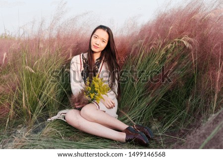 Portrait of beautiful young Chinese woman wearing white sweater dress posing with yellow wild flowers in hand in the pink hairawn muhly field.