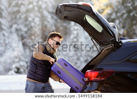 Handsome man going to vacations, loading his suitcase in car trunk. Winter automobile trip in the countryside. Vacation in mountains. Alps, Bavaria, Germany (Deutschland) 