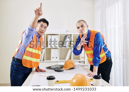 Multi-ethnic team of construction engineers leaning over table with building blueprint and pointing away at something interesting