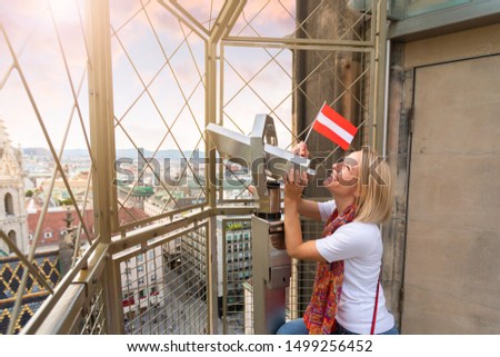 Young woman tourist with the flag of Austria in her hands looks through observation binoculars and enjoys the panorama of the city at sunset in Vienna, Austria