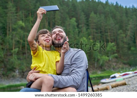 Father and son take pictures of themselves on a smartphone, show grimaces and stick their tongue out onto the camera. Tourists by the river on a summer day