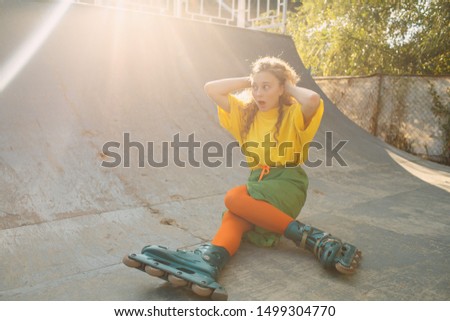 Young woman girl in green and yellow clothes and orange stockings with curly hairstyle roller skating fell in skate park