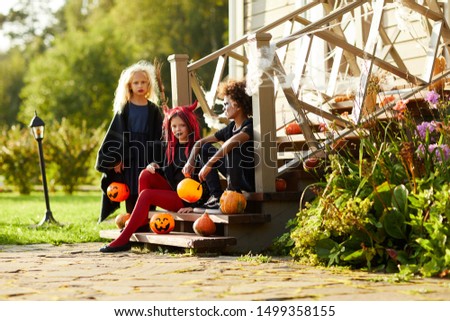 Full length portrait of children trick or treating on Halloween, kids looking at camera while sitting on stairs of decorated house, copy space