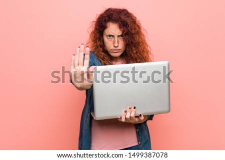 Young caucasian redhead woman holding a laptop standing with outstretched hand showing stop sign, preventing you.