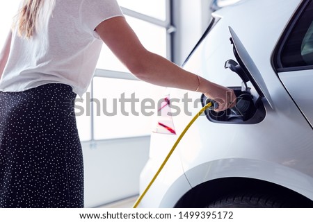 Close Up Of Woman Charging Electric Vehicle With Cable In Garage At Home Royalty-Free Stock Photo #1499395172