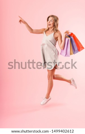 Full length image of caucasian young woman wearing dress looking at copyspace and carrying colorful paper shopping bags isolated over pink background