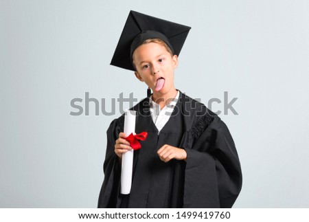 Little boy graduating showing tongue at the camera having funny look on grey background