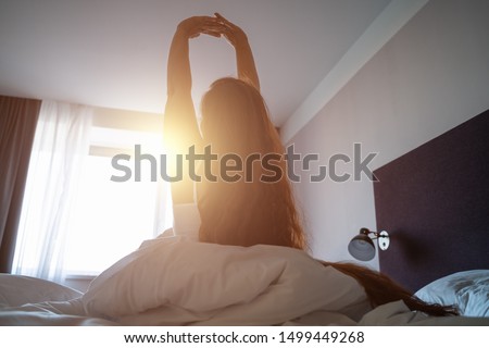 Young woman wakes up, gets up on bed and stretches hands up in morning sunlight, toned Royalty-Free Stock Photo #1499449268