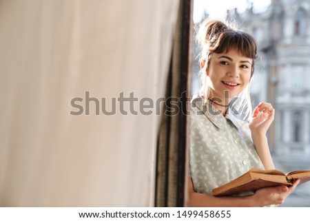 Picture of a cheerful positive smiling cute young beautiful woman reading book on a balcony.
