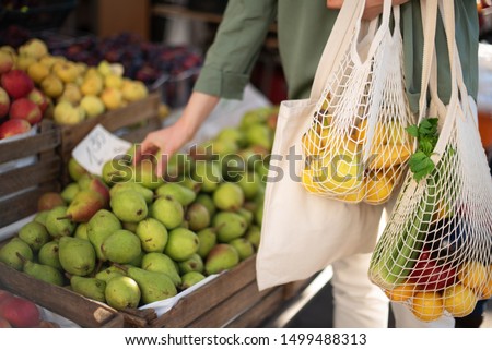 Woman chooses fruits and vegetables at farmers market. Zero waste, plastic free concept. Sustainable lifestyle. Reusable cotton and mesh eco bags for shopping Royalty-Free Stock Photo #1499488313