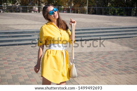 Beautiful woman, has yellow dress, blue sunglasses, leather handbag and belt, enjoy summer view. Urban city portrait. People happy concept. Portrait fashion street style. 