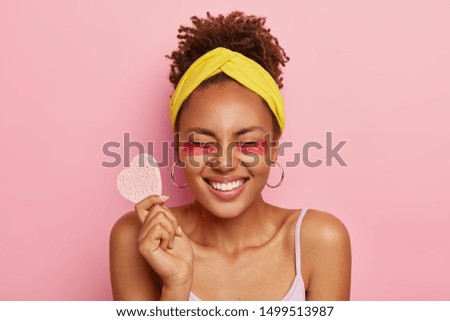 Overjoyed dark skinned woman closes eyes, wears facial patches under eyes, feels relieved and satisfied, holds cosmetic sponge in form of heart, models against pink background. Natural beauty