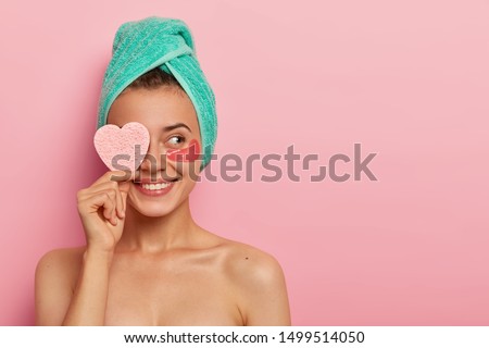 Cheerful young female model has pleasant smile, covers eye with cosmetic sponge, enjoys all benefits of patches, reduces wrinkles, wears wrapped towel on head, has skincare routine after awakening Royalty-Free Stock Photo #1499514050