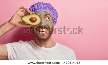 Horizontal view of handsome man covers eye with slice of avocado, smiles happily, applies cosmetic clay mud mask, wears purple showercap, casual white t shirt, models against pink background Royalty-Free Stock Photo #1499514116
