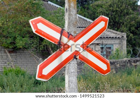 Traffic sign a white cross with a red outline pinned to a pillar against the background of an old house. Warning Sign - 1.3.1 Single Track Railway. Summer season, old rusted road sign.
