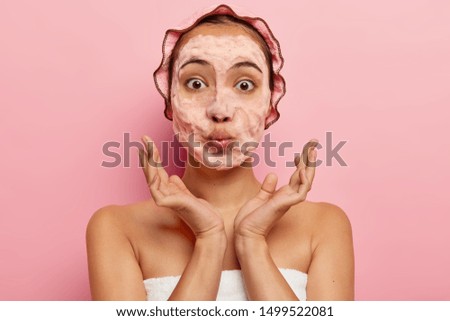 Young shower woman with surprised face expression, washes face with soap, has foam in skin, keeps lips folded, spreads palms, wears showercap, towel around body, isolated over pink background Royalty-Free Stock Photo #1499522081