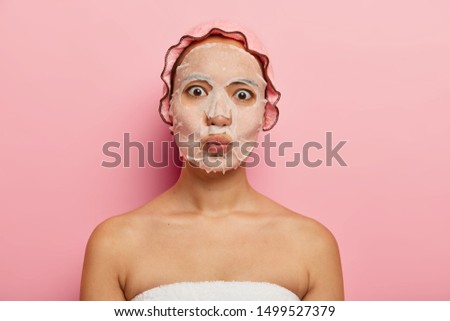 Surprised Chinese woman keeps lips folded, makes grimace, wears paper facial mask for refreshing, has healthy complexion, smooth perfect skin, wears showercap, wrapped in towel after taking bath Royalty-Free Stock Photo #1499527379