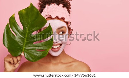 Cropped shot of happy young woman applies facial mud mask on face for removing wrinkless or fine lines, takes bath at bathroom, wears protective rosy headband, holds big plant leaf covers eyes with it Royalty-Free Stock Photo #1499561681